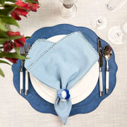 Blue scalloped faux leather placemat with a light blue linen pom-pom napkin, blue glass napkin ring and white plates